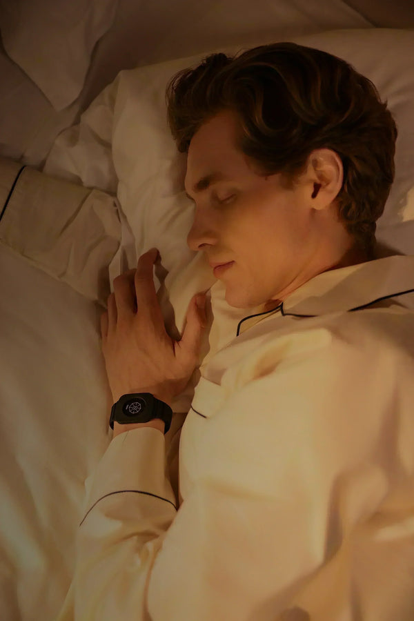 Mann liegt schlafend im Bett bei warmem Licht mit geschlossenem Augen und trägt Eloop Band mit Calm Chip am Handgelenk