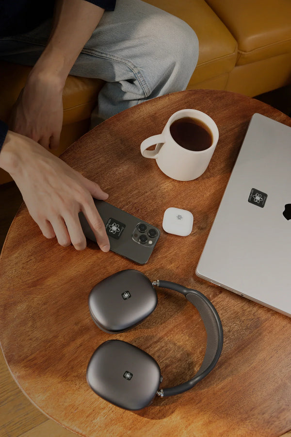 Hand auf Smartphone mit sichtbaren EMF Schutzaufklebern auf Holztisch neben Laptop und Kopfhörern mit EMF Schutzaufklebern sowie Kaffeetasse