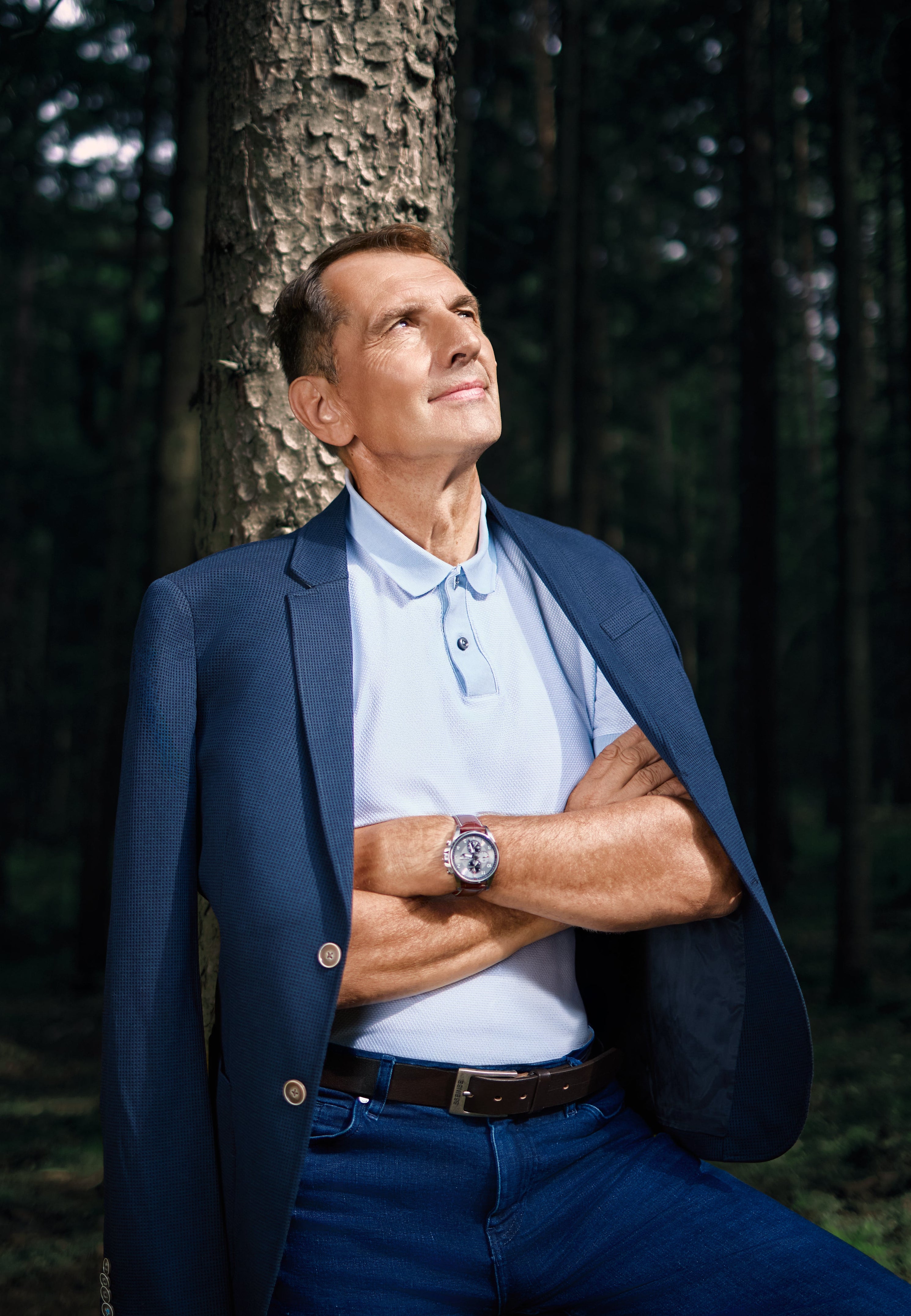 Mann lehnt an Baum im Wald mit verschränkten Armen und blickt nach oben bei Tageslicht