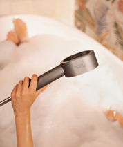 Hand hält den AERO Shower Duschkopf über einer mit Schaum gefüllten Badewanne, Armatur und Wasseroberfläche im Hintergrund