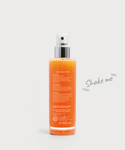 Ageless Body Spray von hinten: transparente Sprühflasche mit silbernem Pumpkopf, gefüllt mit orangefarbener Flüssigkeit, Rückseitenbeschriftung sichtbar, daneben der Hinweis „Shake me“, vor hellem Hintergrund.