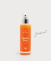 Ageless Body Spray: transparente Sprühflasche mit silbernem Pumpkopf, gefüllt mit orangefarbener Flüssigkeit, Frontansicht mit Produktname „Ageless Body Spray“ und dem Hinweis „Shake me“ daneben, vor hellem Hintergrund.