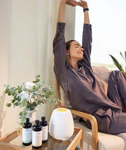 Frau sitzt auf einem Sessel im Innenraum und streckt die Arme, auf dem Beistelltisch stehen ein weißer ProEnergetic Aroma Diffuser und drei Flaschen Duftessenzen neben einer Vase