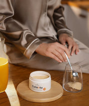 Person in beigefarbener Loungewear gibt mit einem Löffel CollaVita-Pulver aus einer offenen Dose in ein Glas. Die Dose steht auf einem runden Holztablett, daneben eine Karaffe mit Getränk, alles in warmer, natürlicher Wohnatmosphäre.