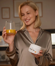 Lächelnde Person in seidiger Loungewear hält ein Glas mit orangem Getränk und eine Dose CollaVita in den Händen, in einer warmen, wohnlichen Küchenumgebung.