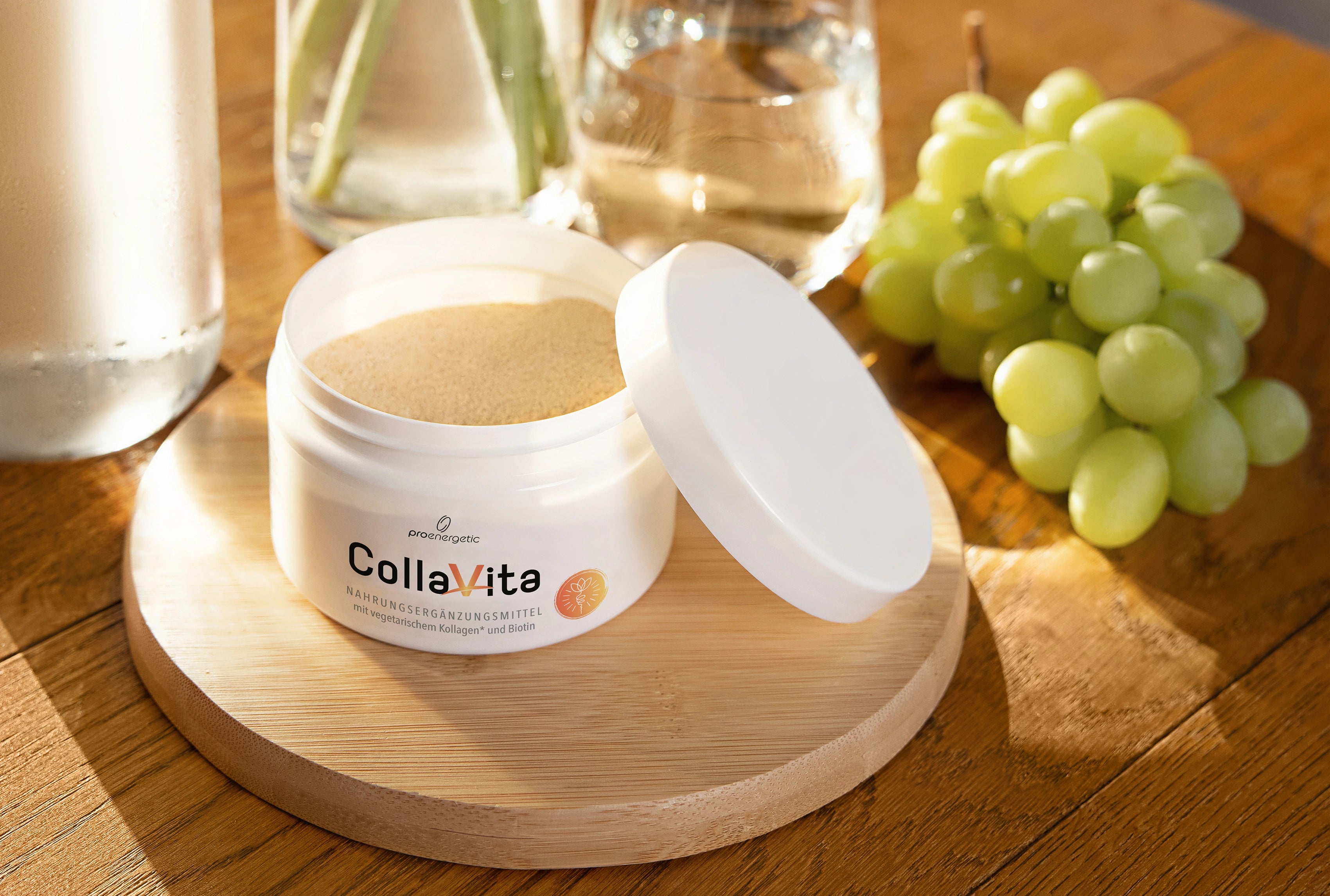Geöffnete Dose CollaVita mit Pulver auf einem Holztablett; daneben liegen grüne Trauben, im Hintergrund stehen Wasserglas und Karaffe.