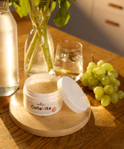 Offene CollaVita-Dose mit Pulver steht auf einem Holztablett auf einem Tisch, daneben ein Glas Wasser, eine Karaffe, grüne Trauben und eine Vase mit Blumen in warmem Tageslicht.