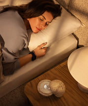 Dormea on its natural sand stone on a bedside table beside a woman sleeping peacefully in bed.