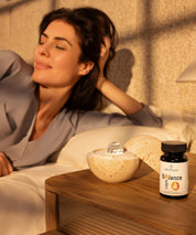 Woman relaxing in bed beside Dormea and ProEnergetic Balance Caps supplement on a bedside table.