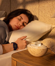 Woman sleeping peacefully while wearing the Eloop Band with sleep Chip, with the Dormea natural sand stone standing on the bedside table.