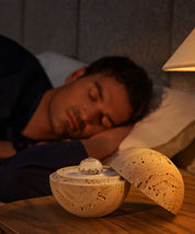 Dormea on its sand stone on a bedside table, with a man sleeping peacefully in the background.