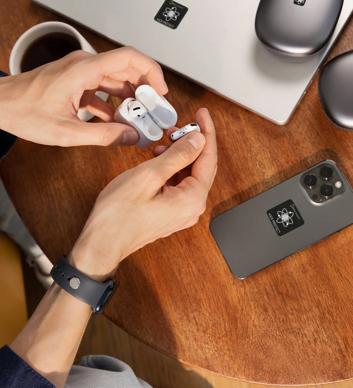 Hände nehmen kabellose In-Ear-Kopfhörer aus einem Ladecase. Auf dem Holztisch liegen Smartphone und Laptop, beide mit E-Protect-Audio-Aufklebern versehen, daneben eine Kaffeetasse.