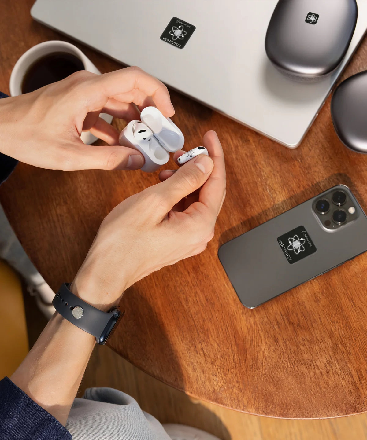 Hände nehmen kabellose In-Ear-Kopfhörer aus einem Ladecase. Auf dem Holztisch liegen Smartphone und Laptop, beide mit E-Protect-Audio-Aufklebern versehen, daneben eine Kaffeetasse.