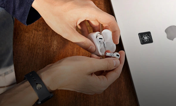 Mann hält AirPod über Ladecase auf Tisch mit Laptop und Smartphone mit EMF Schutzaufklebern sowie Smartwatch mit EMF Schutzpin