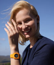 Lächelnde Person im Freien mit blonden Haaren, die ein oranges Eloop Band mit eingesetztem Eloop Chip Flow am Handgelenk trägt.