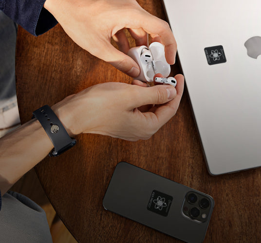 Mann hält AirPod über Ladecase auf Tisch mit Laptop und Smartphone mit EMF Schutzaufklebern sowie Smartwatch mit EMF Schutzpin