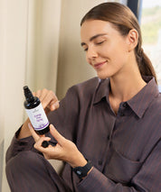 Person sitzt im Innenraum und hält das Energy Flow Spray in schwarzer Sprühflasche, betrachtet das Etikett bei natürlichem Licht