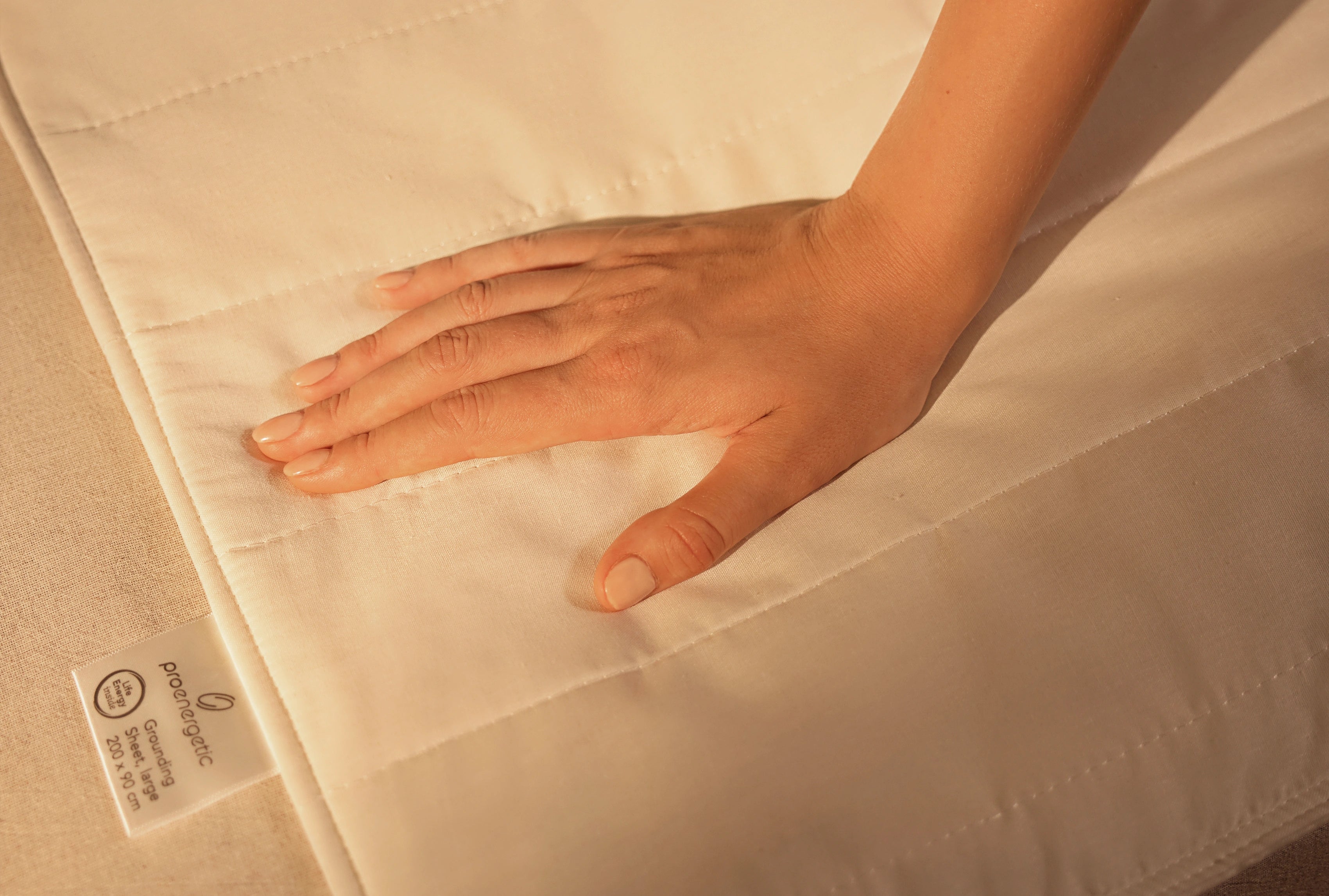 Hand liegt auf der weißen Grounding Sleep Mat mit gesteppter Oberfläche, Textiletikett am Rand sichtbar