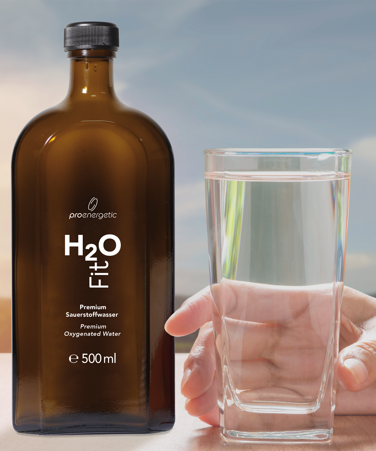 Braune Glasflasche H2O Fit von ProEnergetic neben gefülltem Wasserglas in einer Hand vor hellem Hintergrund.
