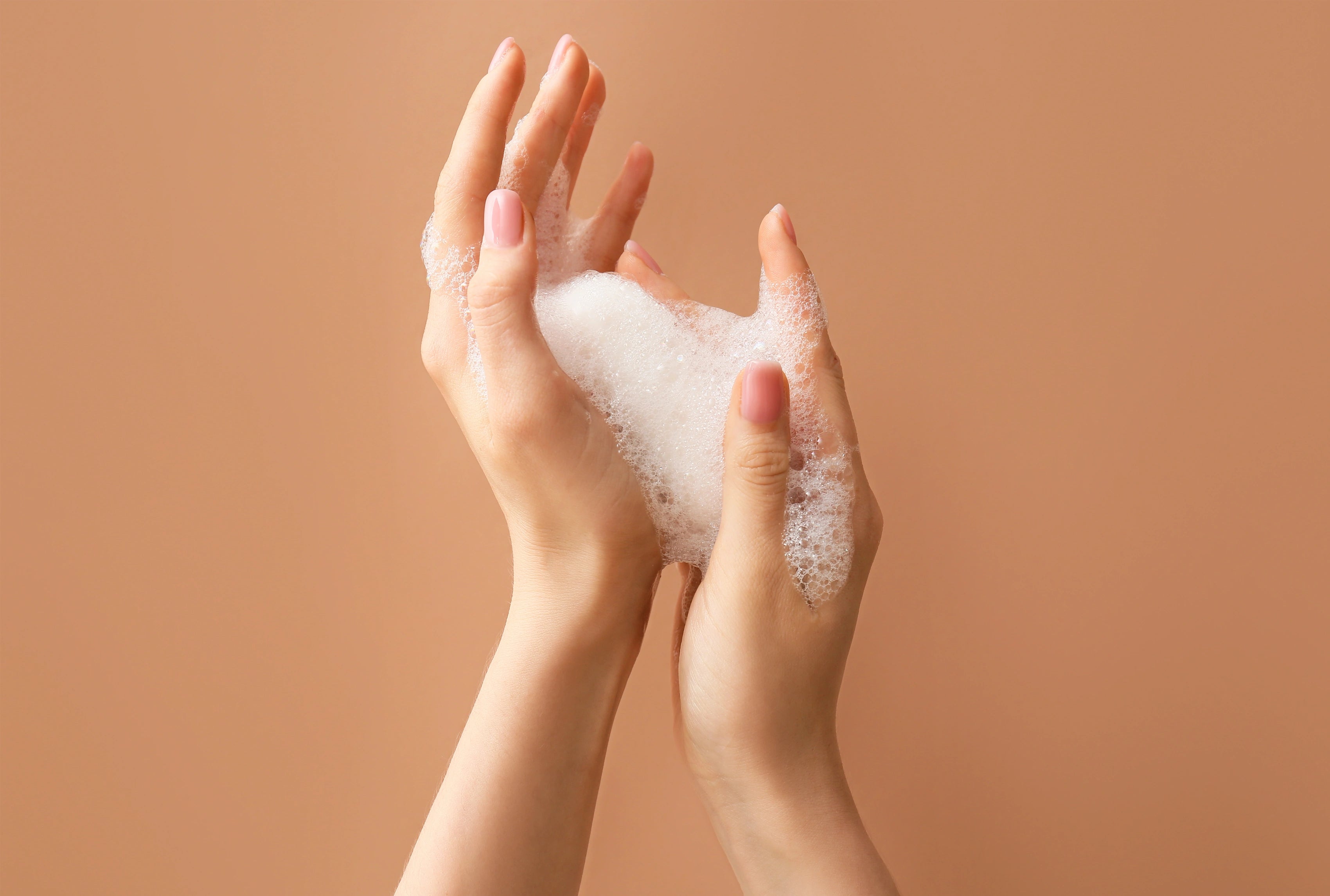 Zwei gepflegte Hände, die einen luftigen, cremigen Schaum – passend zum Happy Shower Foam – sanft zwischen den Fingern verteilen, vor warmem, beigefarbenem Hintergrund.