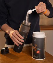 Eine Person gibt mit einem Messlöffel Pulver aus der ISOmove Fit Dose von proenergetic in eine schwarze Trinkflasche. Die geöffnete Dose steht auf einem Holztisch, warmes Innenlicht, Fokus auf Zubereitung des Mineral-Vitamin-Drinks.