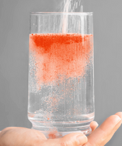 Ein Glas Wasser wird gehalten, während ISOmove Fit-Pulver hineingestreut wird. Das orangefarbene Pulver löst sich sprudelnd auf und färbt das Wasser gleichmäßig – symbolisiert schnelle Löslichkeit, Frische und den Blutorangen-Geschmack.
