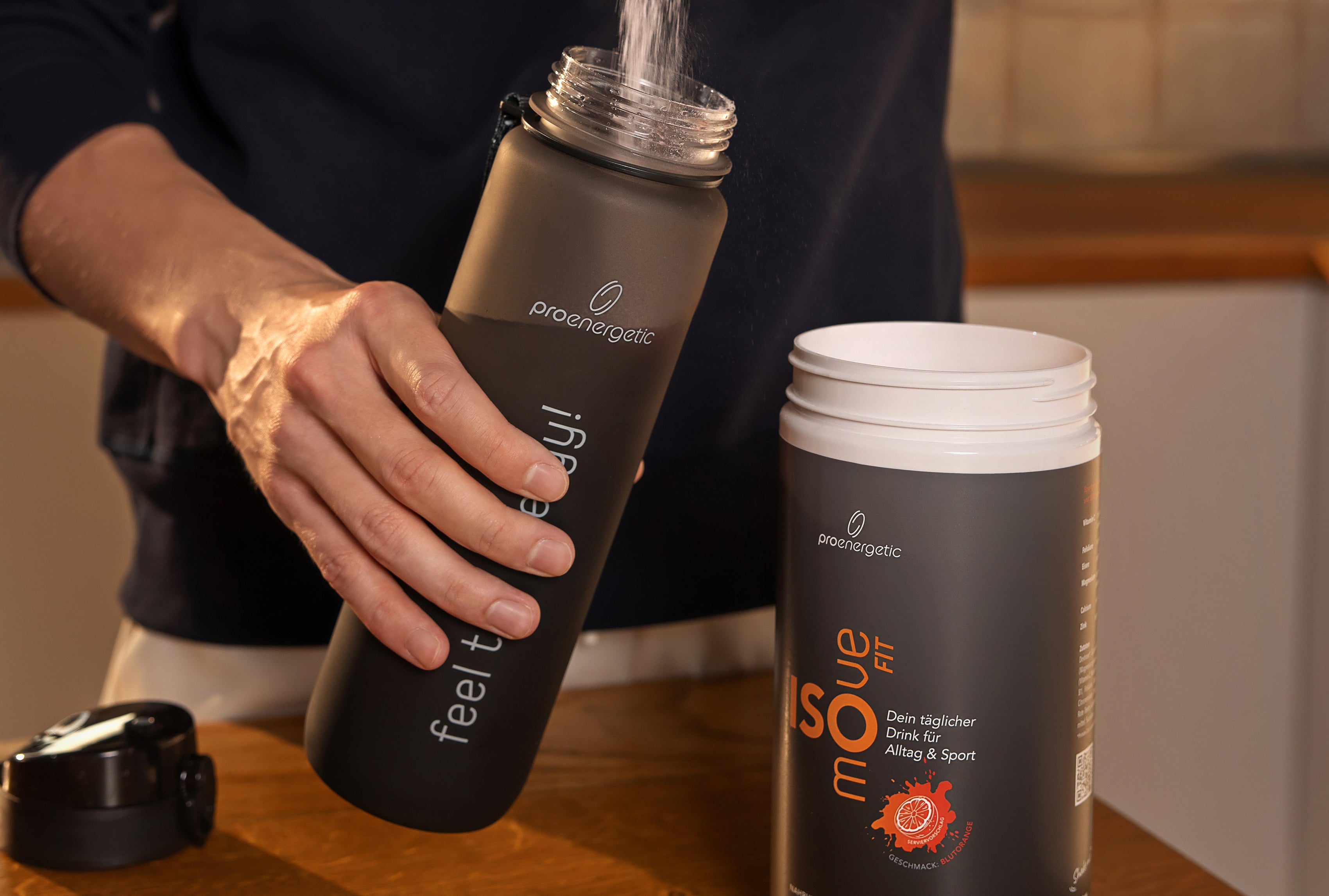 Eine Person hält eine dunkle Trinkflasche und gießt Wasser hinein. Daneben steht eine geöffnete Dose ISOmove Fit auf einem Holztisch. Die Szene zeigt die Zubereitung des Mineral-Vitamin-Drinks.
