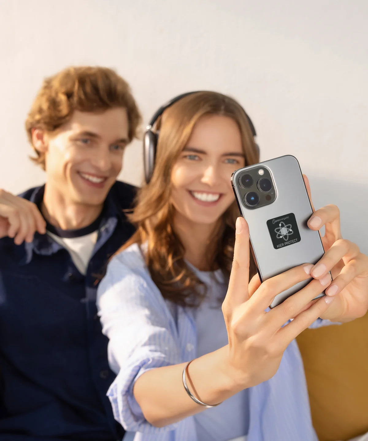 Zwei Personen sitzen im Innenraum, eine Frau hält ein Smartphone mit angebrachtem KIER Protect Mobile Element vor sich und lächelt