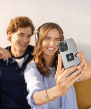 Zwei Personen sitzen im Innenraum, eine Frau hält ein Smartphone mit angebrachtem KIER Protect Mobile Element vor sich und lächelt