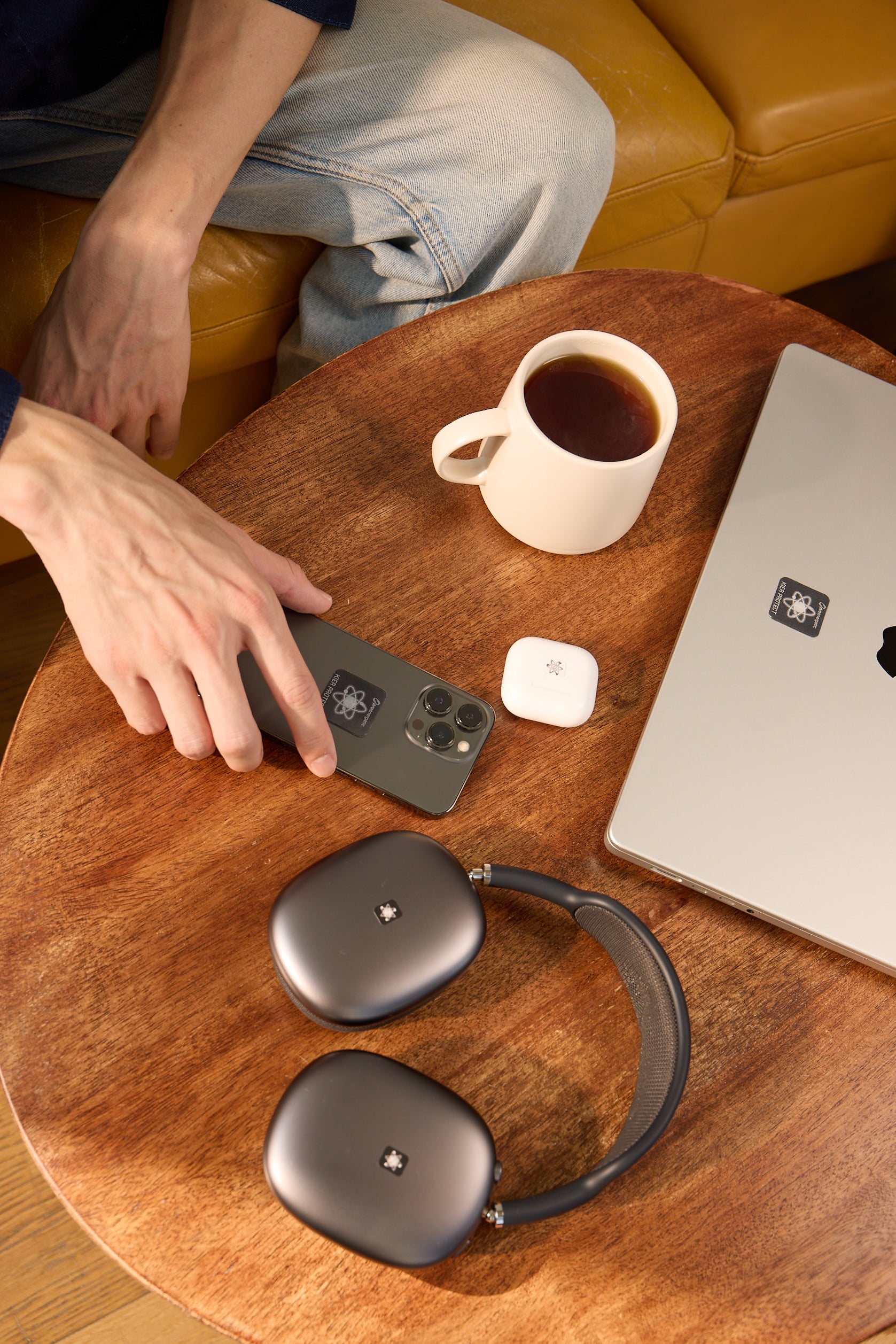 Hand auf Smartphone mit sichtbaren EMF Schutzaufklebern auf Holztisch neben Laptop und Kopfhörern mit EMF Schutzaufklebern sowie Kaffeetasse