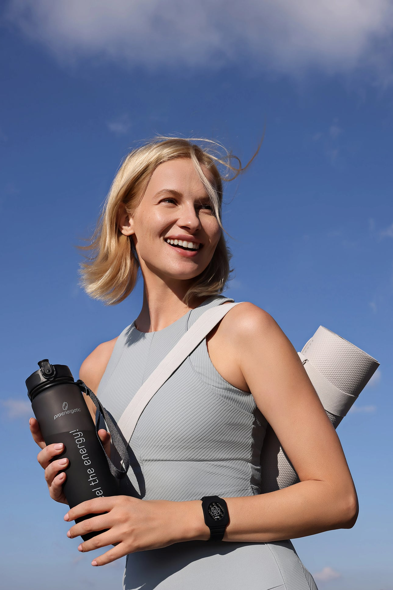 Lächelnde Frau mit umgehängter Yogamatte und schwarzer Pure Move Trinkflasche vor blauem Himmel