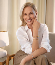 Frau sitzt in einem Innenraum auf einem Stuhl und trägt den Lunara Cuff Armreif in Gold und Silber an den Unterarmen