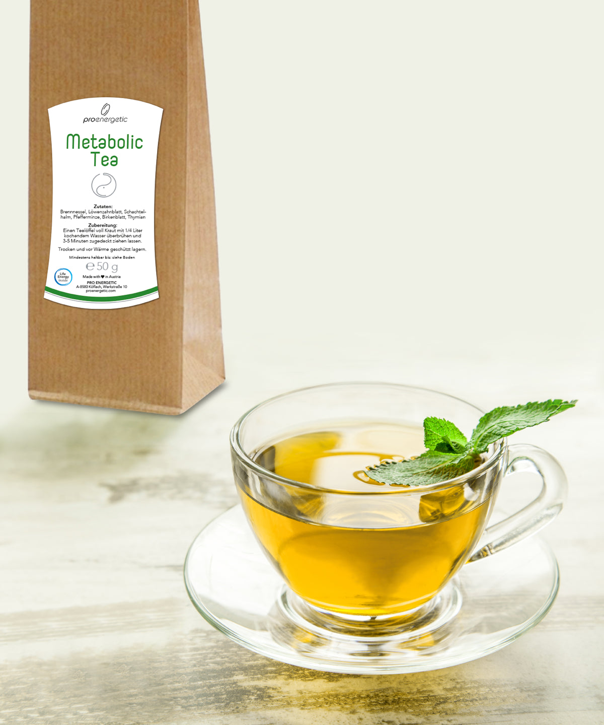 Gläserne Teetasse mit aufgebrühtem Metabolic Tea und Minzblatt auf Untertasse, braune Teeverpackung im Hintergrund.