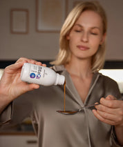 Frau in Satin-Pyjama gießt MitoQi Liquid von proenergetic aus der Flasche auf einen Löffel; Anwendung eines veganen Nahrungsergänzungsmittels zur Unterstützung des Energiestoffwechsels in einer warmen Wohnküchen-Umgebung.