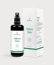 Dunkle Sprühflasche Nature Care Essence neben weißer Produktverpackung auf hellem Hintergrund.