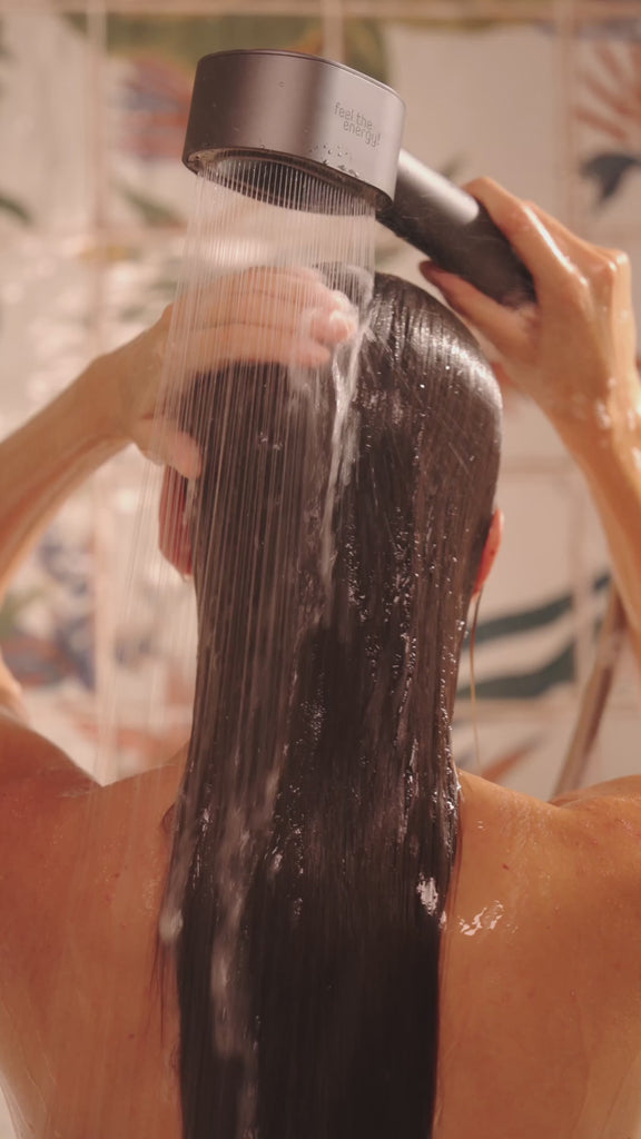 Person duscht von hinten gesehen und hält den AERO Shower Duschkopf über nassem Haar, Wasserstrahlen fließen gleichmäßig herab, benutzt beide Aufsätze nacheinander