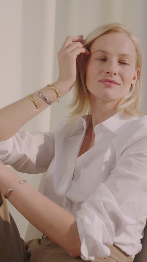 Frau sitzt in einem Innenraum auf einem Stuhl, lächelt und trägt die Lunara Cuff Armreifen in Gold und Silber mehrfach am Unterarm und Handgelenk