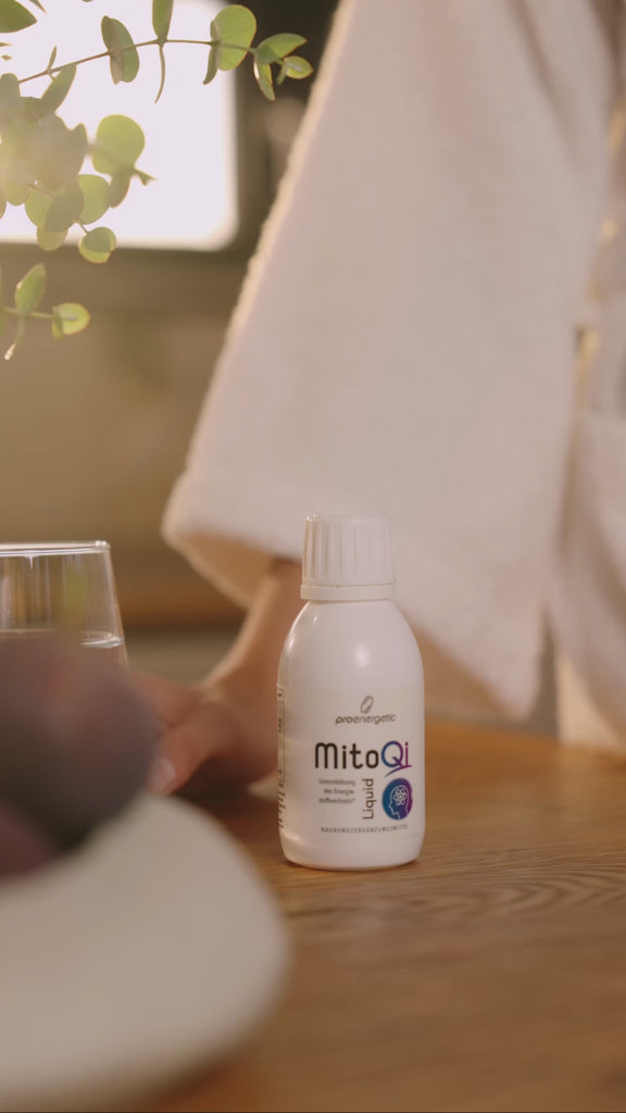 MitoQi Liquid von proenergetic: Person im hellen Bademantel gießt das braune, flüssige Supplement aus einer weißen MitoQi-Flasche in einen Löffel. Nimmt es zu sich. Natürliche, warme Lichtsituation mit Pflanzendetail im Hintergrund. Video.