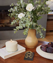 Seed of Happiness Goodie als goldenes Ornament auf schwarzem Sockel, platziert auf einem Holztisch neben Kerze, Vase und Obstschale