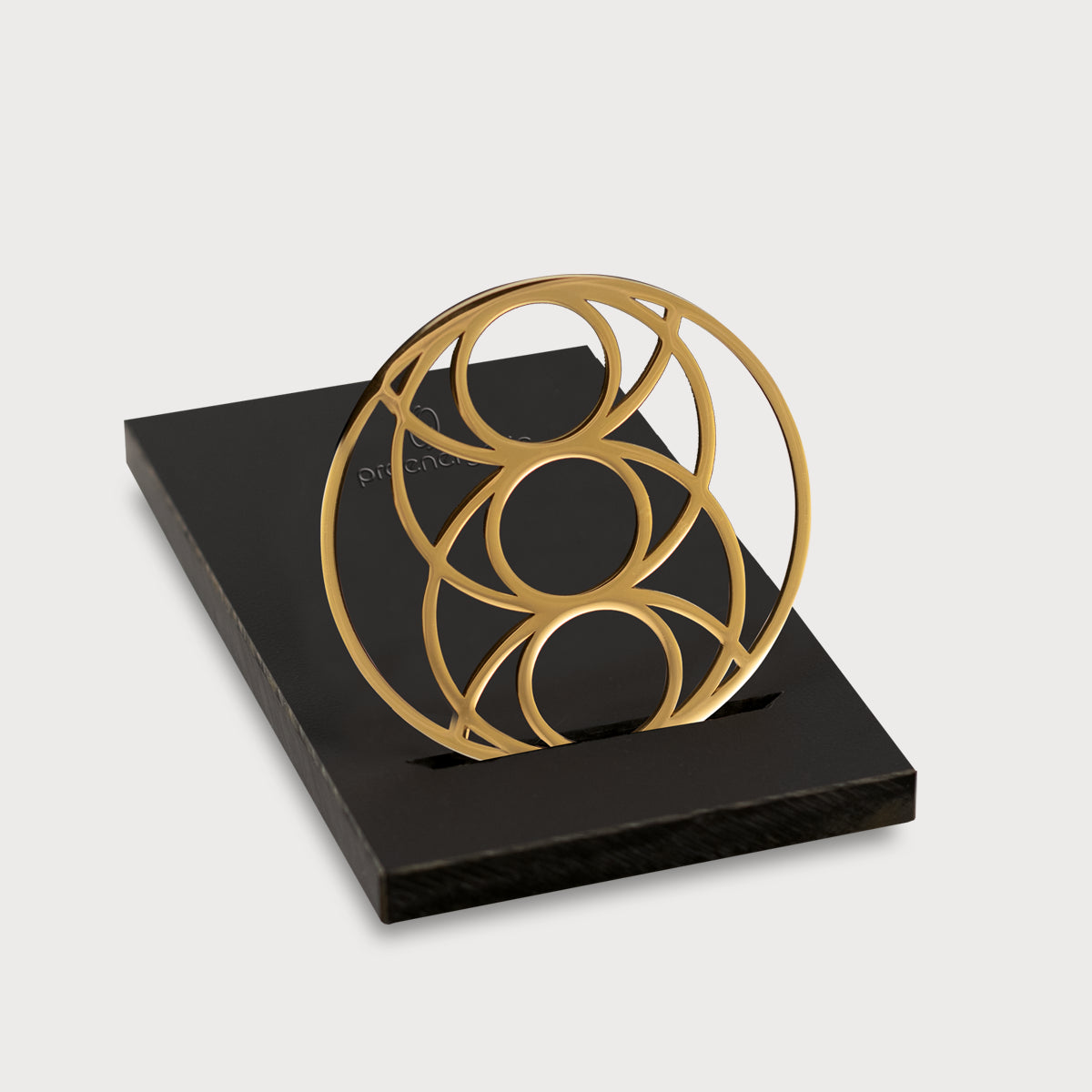 Seed of Happiness Goodie als goldenes Ornament mit kreisförmigem Design auf schwarzem Sockel vor hellem Hintergrund