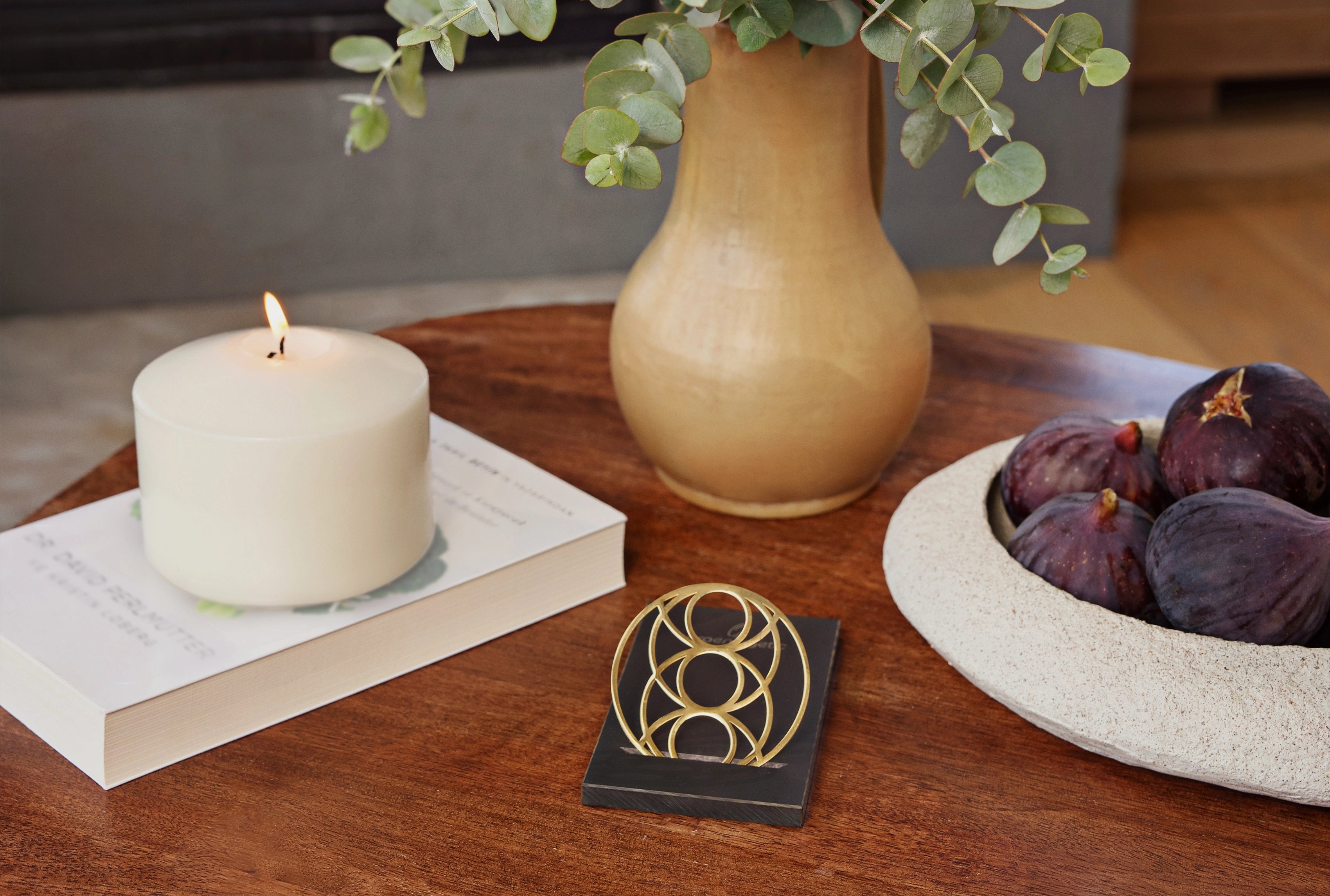 Seed of Happiness Goodie als goldenes Ornament auf schwarzem Sockel, platziert auf einem Holztisch neben Kerze, Vase und Obstschale