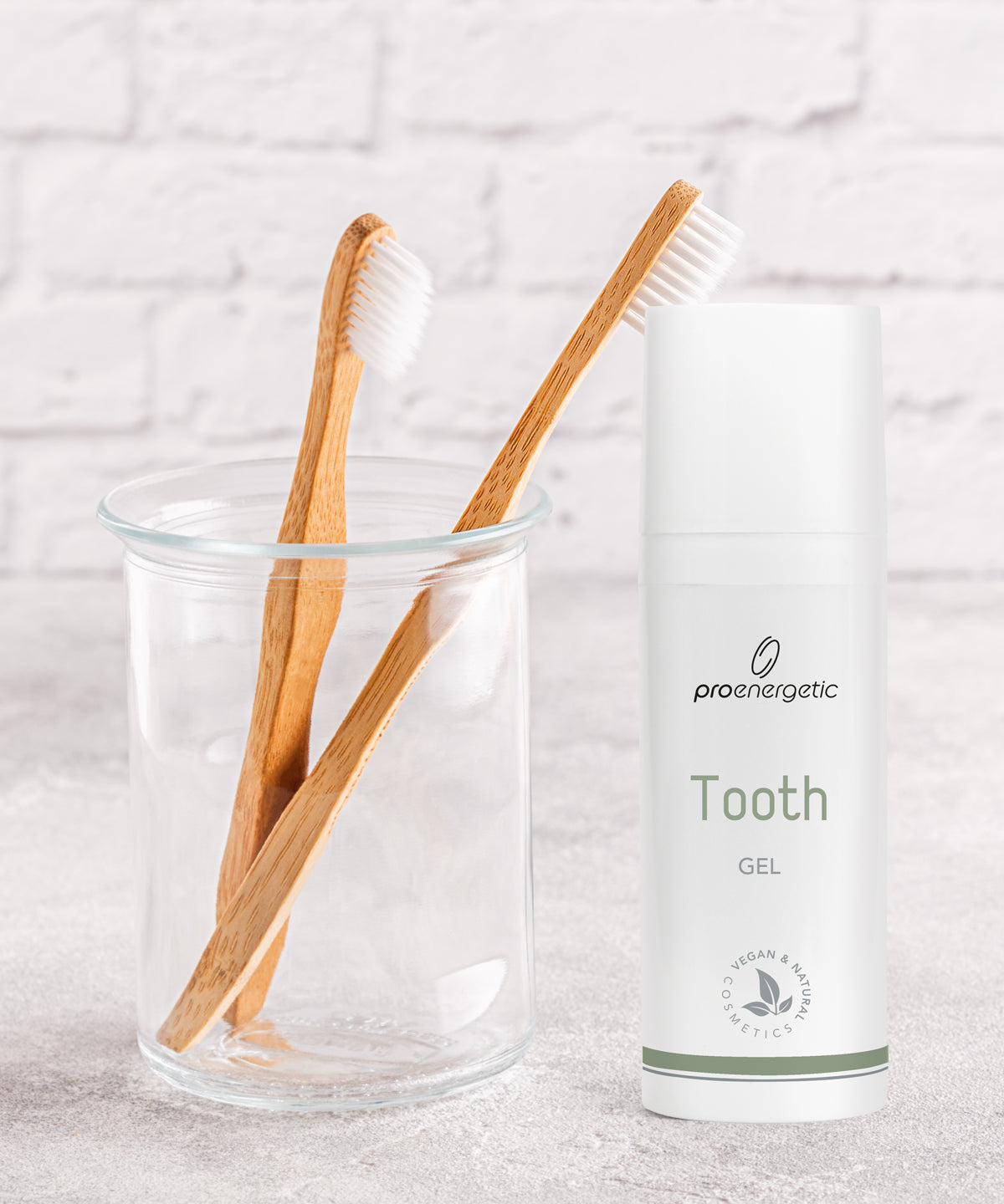 Zwei Bambuszahnbürsten in Glasbecher neben ProEnergetic Tooth Gel Spender vor weißer Ziegelwand.
