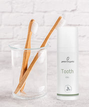 Zwei Bambuszahnbürsten in Glasbecher neben ProEnergetic Tooth Gel Spender vor weißer Ziegelwand.