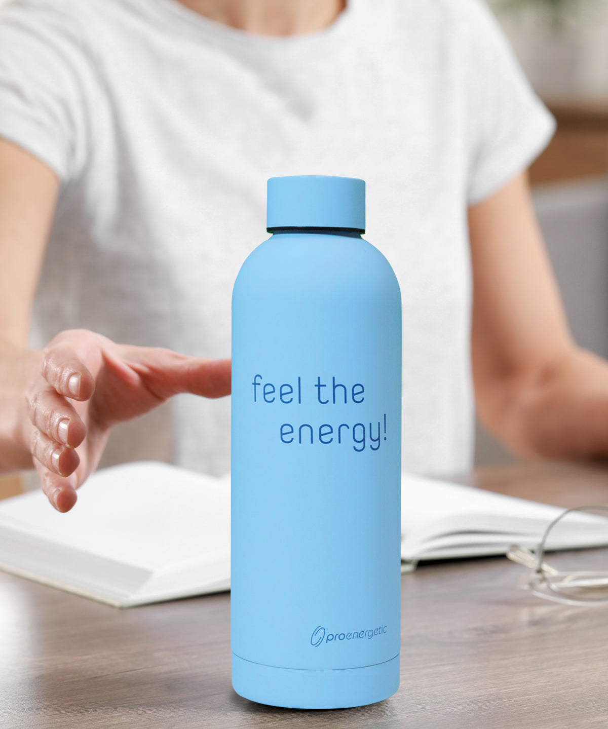 Vita Bottle Trinkflasche in hellblauer Ausführung steht auf einem Tisch vor einer Person mit geöffnetem Buch im Hintergrund